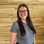 Portrait of Magdalena Ottinger with glasses and long brown hair, wearing a gray polo shirt, smiling in front of a wooden wall.