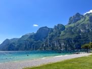Kiesstrand am Gardasee in Riva del Garda mit türkisfarbenem Wasser, Schwimmern und dramatischen Bergen unter blauem Himmel.