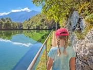 Frau mit rosa Kappe und Zöpfen blickt auf den Plužna-Stausee im Soča-Tal. Klares türkisfarbenes Wasser spiegelt Berge und Himmel.