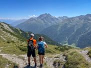 Zwei Wanderer mit Rucksäcken stehen auf einem Bergweg in den Allgäuer Alpen und betrachten ein Panorama von Gipfeln und Tälern unter blauem Himmel.