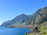 Panoramablick auf den Gardasee mit steilen Bergen hinter einer Uferstadt. Blaues Wasser im Vordergrund, terrassierte Gebäude an Hängen.