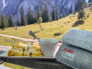 Plate of Kaiserschmarrn with powdered sugar and a cup of berry sauce on a wooden table. Eurohike hat beside it, alpine meadow and mountains behind.