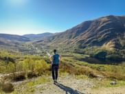 Wanderin mit Rucksack blickt auf das Kinlochleven-Tal am West Highland Way. Berge, See und Dorf unter blauem Himmel sichtbar.