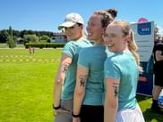Three smiling women in turquoise shirts with matching temporary tattoos on their arms at Grabenseelauf 2026 sports event on a sunny day.