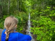 Woman with braided hair in blue jacket viewing a narrow stream flowing through lush green forest near Sautens on the E5 Alpine crossing route.