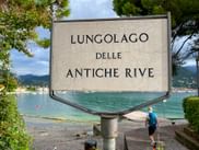 Weißes Schild mit der Aufschrift 'Lungolago delle Antiche Rive' an der Promenade mit dem türkisfarbenen Wasser des Gardasees und Bergen im Hintergrund.