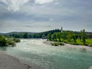 Turquoise Isonzo River with rocky banks flowing through Sagrado village in Friuli. Church spire visible among green trees and hills.