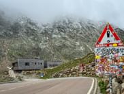 Road at Timmelsjoch Pass with colorful sticker-covered sign and mountain building. Snow-dusted rocky peaks visible in misty background.