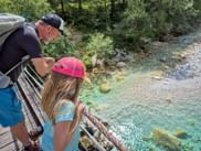 Wanderer mit Rucksack und Mädchen mit rosa Kappe stehen auf Holzbrücke und blicken auf den kristallklaren türkisfarbenen Soča-Fluss.