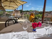 Set table with wine glasses and pink roses on a terrace overlooking Barolo village in Piedmont, with historic buildings and vineyards.