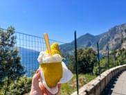 Hand hält zitronenförmiges Eis mit Sahne in Limone sul Garda. Gardasee und Berge im Hintergrund sichtbar.