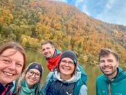 Five smiling people in outdoor jackets taking a selfie by the Danube river with autumn-colored hills in the background during a company trip.