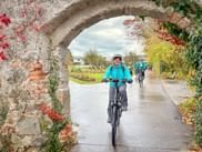 Group of cyclists in turquoise jackets riding through ivy-covered stone archway on paved path during Eurofun Touristik company outing.
