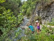 Two hikers with backpacks walking along a rocky stream in Rosengartenschlucht gorge near Imst, surrounded by lush green vegetation and rock walls.