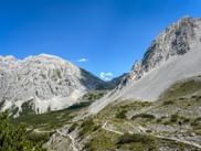 Dramatische Kalksteingipfel des Karwendelgebirges bei Innsbruck mit Geröllhalden, grünen Almwiesen und einem einzelnen Wanderer auf dem Weg.