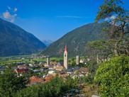 Alpine village of Mals in South Tyrol with church spire, surrounded by green mountains and valley. Blue sky above forested mountain slopes.