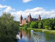 Johannisburg Castle with red sandstone towers rises above green trees beside a calm river. A small boat travels on the water under blue sky.