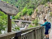Hiker with backpack on wooden bridge at Wellerbrücke in Ötztal. Wooden sign visible, rocky gorge with rushing water and lush forest in background.