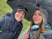 Two smiling female hikers sharing a black umbrella on a rainy day in Wachau. One wears a rain jacket and cap, the other a rain poncho.