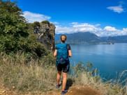 Wanderin mit blauem Rucksack steht auf Felsen an der Rocca di Manerba mit Blick auf den Gardasee, Bergen und blauem Himmel im Hintergrund.