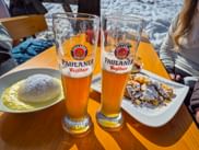 Zwei Paulaner Weißbier-Gläser mit österreichischen Desserts: Germknödel mit Vanillesauce und Kaiserschmarrn mit Puderzucker auf gelbem Tisch.