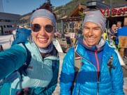 Zwei lächelnde Frauen in blauer Skitourenausrüstung mit Rucksäcken und Skiern machen ein Selfie in einem Bergresort beim Wintersporttag 2026.