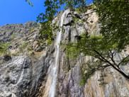 Hoher Wasserfall, der eine Felswand am Sentiero del Sole am Gardasee hinabfließt, eingerahmt von grünen Ästen vor blauem Himmel.