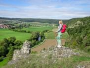 Hiker with red backpack standing on rocky outcrop overlooking green valley in Altmühltal with villages, fields, and forested hills.