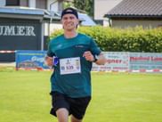 Smiling runner wearing teal shirt and cap with race bib 535 running on grass field at Grabenseelauf 2026, with buildings and hedges in background.