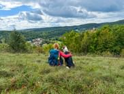 Wanderer mit Rucksack sitzt auf Grashang mit schwarz-weißem Hund und blickt auf Tal mit Dorf und bewaldeten Hügeln bei Pappenheim.