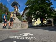 Two hikers with backpacks viewing a historic fountain statue in Imst. Arrow marking on street points toward Schlöcht.