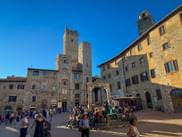 Piazza della Cisterna in San Gimignano mit mittelalterlichem Steinbrunnen, historischen Türmen und Touristen unter klarem blauen Himmel in der Toskana.