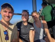 Drei lächelnde Wanderer in einer Valluga Seilbahn-Gondel mit Bergblick durch die Fenster. Blauer Himmel und felsige Gipfel im Hintergrund.