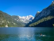 Person swimming in Gosausee lake with snow-capped Dachstein glacier and forested mountains under clear blue sky.