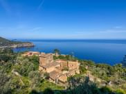 Aerial view of Deia village in Mallorca with traditional stone houses nestled among green vegetation on coastal cliffs above the blue Mediterranean Sea.