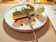 Stück Pistazienkuchen mit Schlagsahne auf weißem Teller, dekoriert mit roten und orangen Saucenpunkten und gehackten Pistazien obenauf.