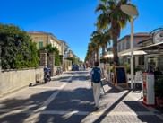 Wanderer mit Rucksack und Wanderstöcken auf einer gepflasterten Straße mit Palmen in Cecina Mare, Toskana. Wohngebäude und Cafés unter blauem Himmel.