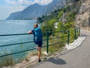 Wanderin mit blauem Rucksack steht am Geländer mit Blick auf den Gardasee bei Limone, Berge und Küste im Hintergrund sichtbar.