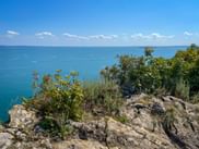 Felsige Klippe mit wilder Vegetation mit Blick auf das türkisfarbene Wasser des Gardasees unter blauem Himmel mit vereinzelten Wolken.