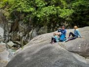 Three hikers in blue clothing sitting on large granite rocks beside a mountain stream in Passer Gorge, surrounded by lush green vegetation.