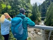 Two hikers with backpacks on a bridge viewing the Passer Gorge, surrounded by dense forest and a mountain stream below.