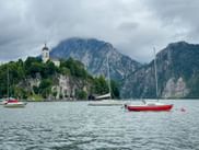 Sailboats moored on lake with Traunkirchen church on rocky peninsula, dramatic alpine mountains in background under cloudy sky.