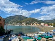Colorful boats with striped covers line the beach in Levanto, Liguria. Mountains rise behind the bay with coastal buildings along the shore.
