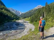 Wanderer mit blauem Rucksack blickt ins Almajurtal mit Fallesinbach, umgeben von bewaldeten Hängen und Felsgipfeln in den Allgäuer Alpen.