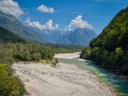 Türkisfarbener Soča-Fluss fließt durch breites Kiesbett im Tal der Julischen Alpen, umgeben von grünen Wäldern und Bergen unter blauem Himmel.