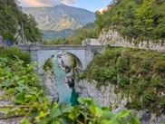 Steinbogenbrücke über eine enge Schlucht mit türkisfarbenem Soča-Fluss, umgeben von grüner Vegetation und den Julischen Alpen.