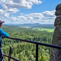 Hiker with blue backpack at Pfaffenstein viewpoint overlooking forested valleys and fields. Distinctive rock formation stands beside the railing.