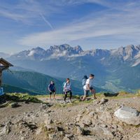 Drei Wanderer neben einem hölzernem Kreuz auf einem steinigen Weg im Naturpark Dolomiten mit Bergpanorama im Hintergrund