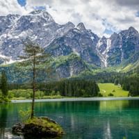 Emerald green lake Laghi di Fusine with small rocky island and lone tree. Snow-capped mountain peaks rise behind dense forests under cloudy sky.