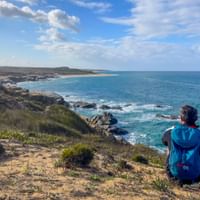 Hiker with blue backpack sitting on coastal cliff overlooking turquoise Atlantic Ocean near Porto Covo, Praia do Sissal, Rota Vicentina, Portugal.